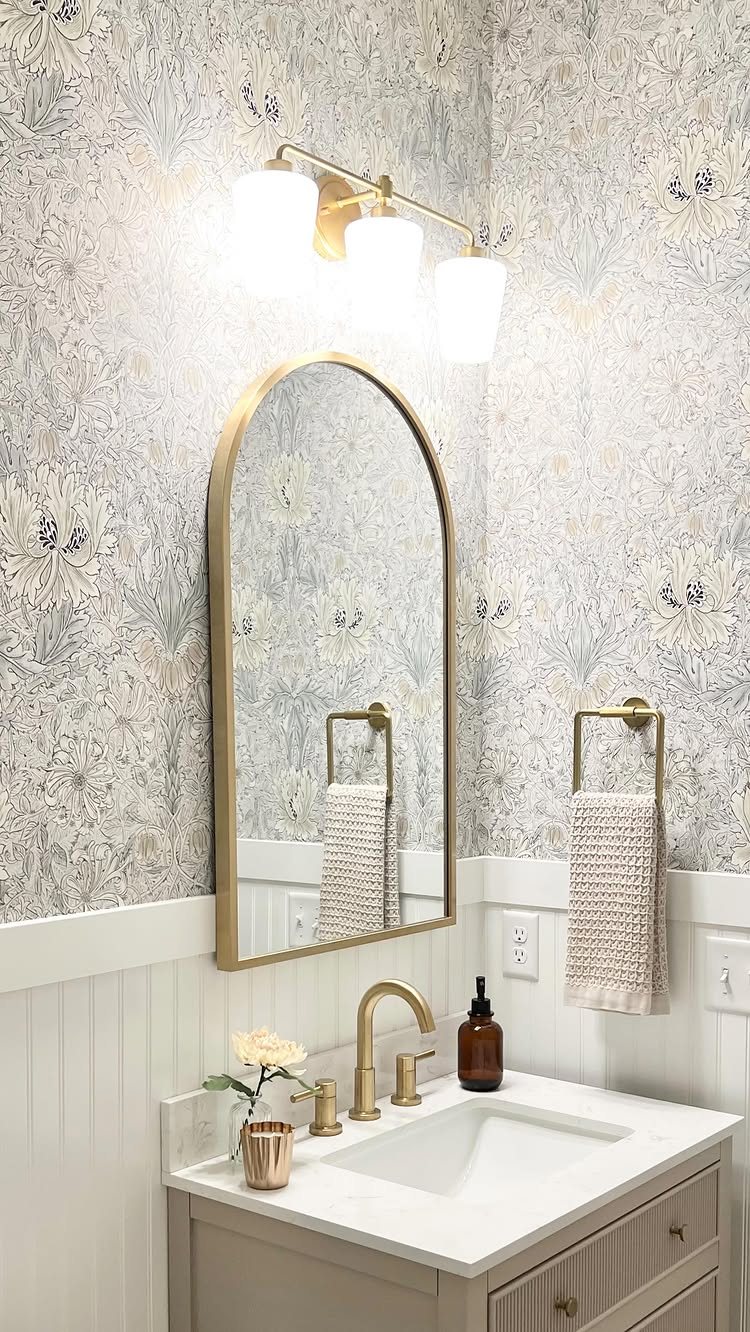 White half-height beadboard with floral wallpaper and gold bathroom fixtures above a white vanity.