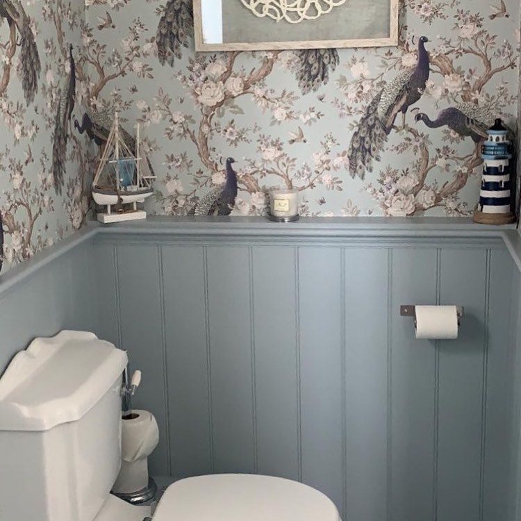 Light blue beadboard wainscoting installed on a bathroom wall beneath peacock patterned wallpaper.