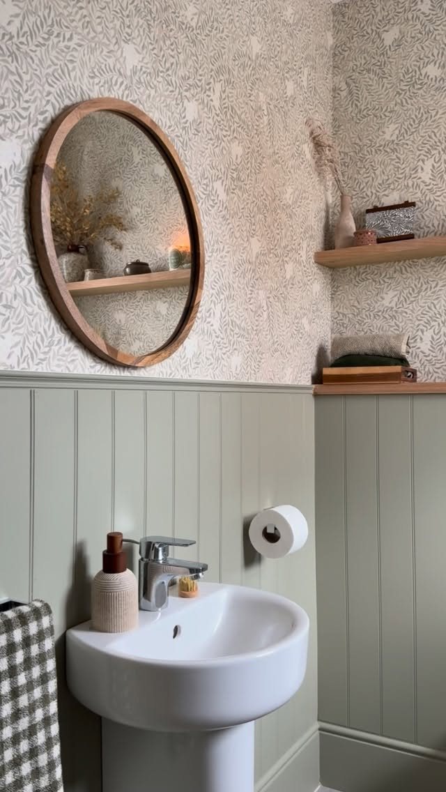 White pedestal sink positioned against sage green beadboard wainscoting and patterned wallpaper in a bathroom.