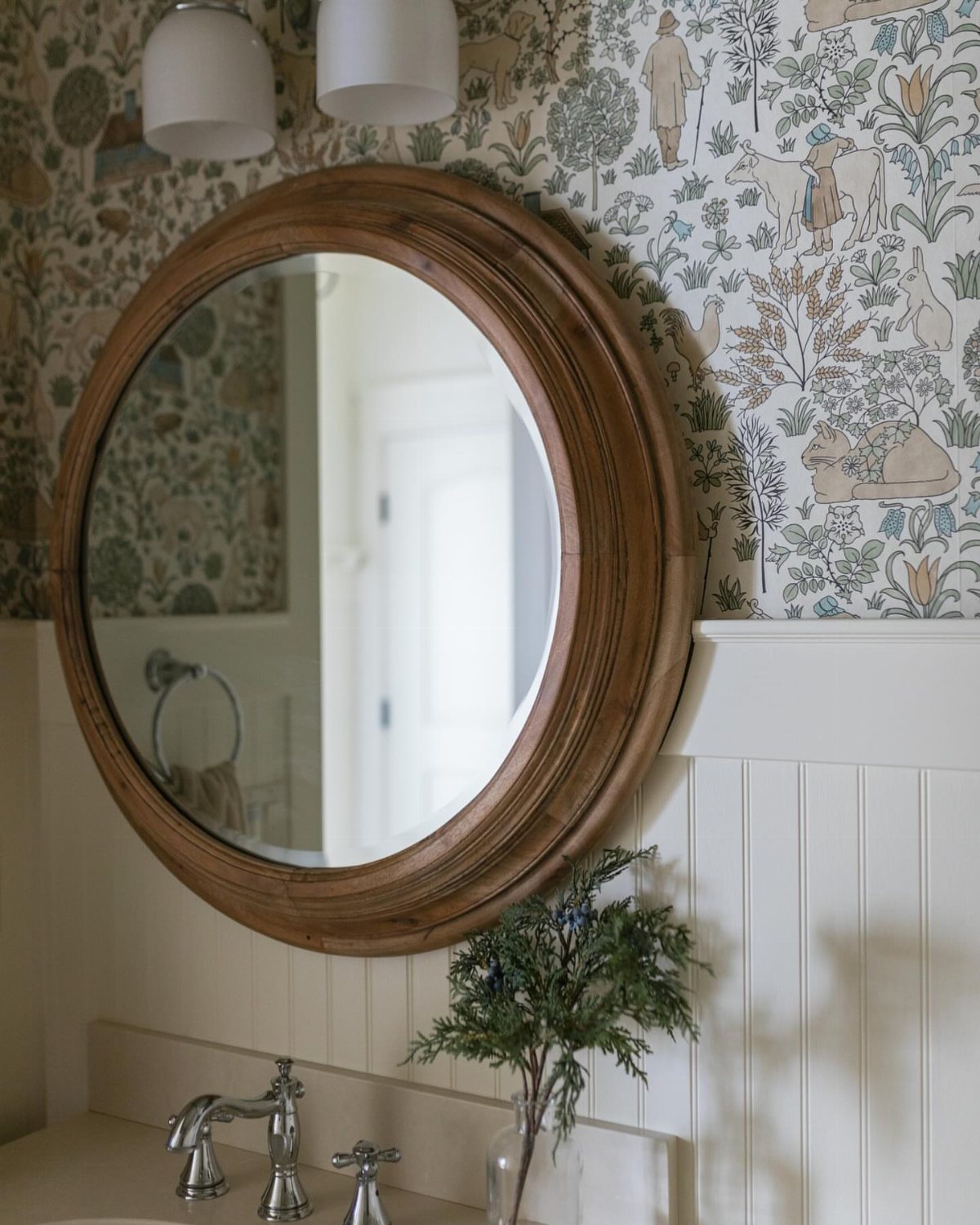 Round wooden mirror hanging on patterned wallpaper above white beadboard wainscoting in a bathroom.