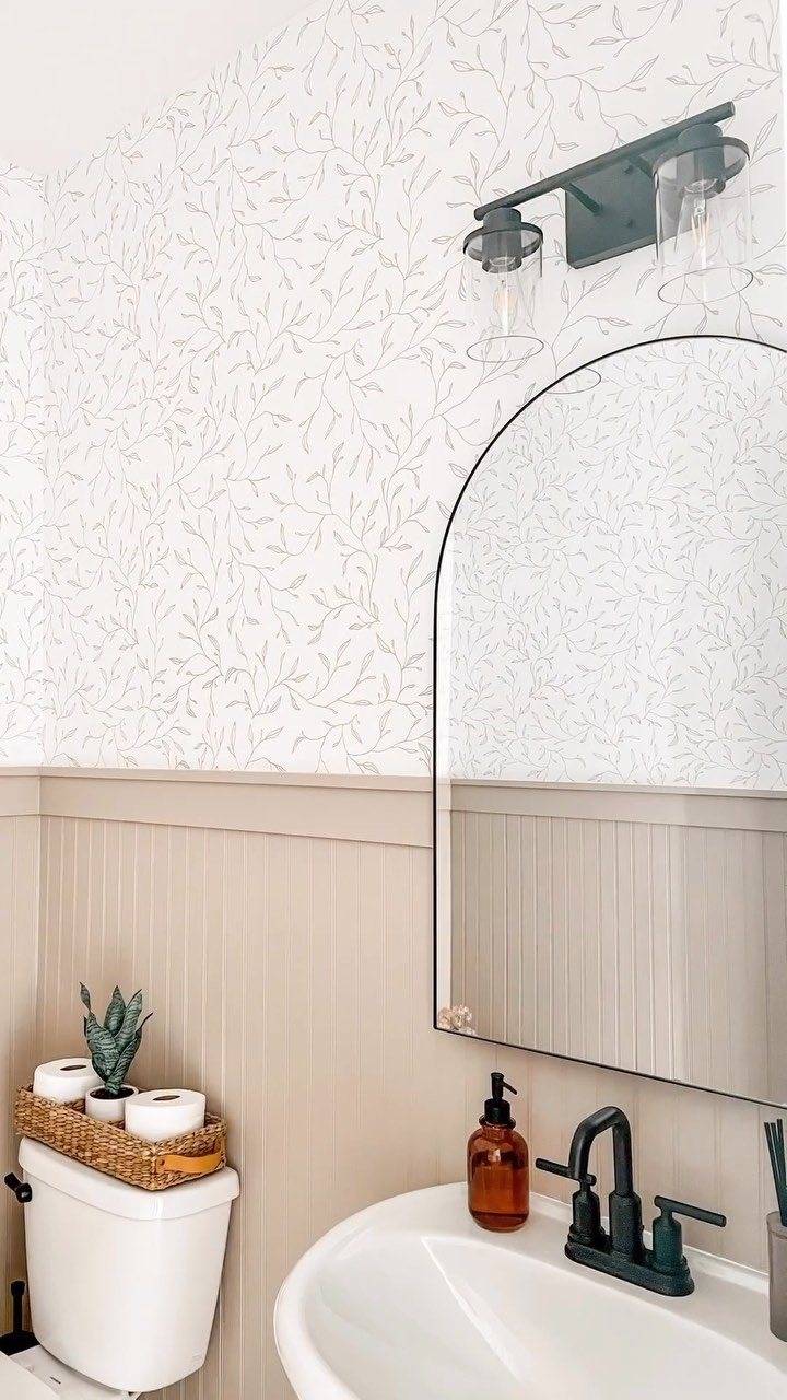 Beige beadboard wainscoting installed on a bathroom wall below white patterned wallpaper.