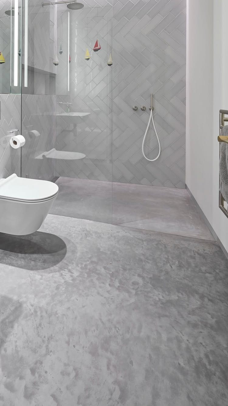 Grey concrete flooring in a modern bathroom with herringbone tiled walls and a white toilet.