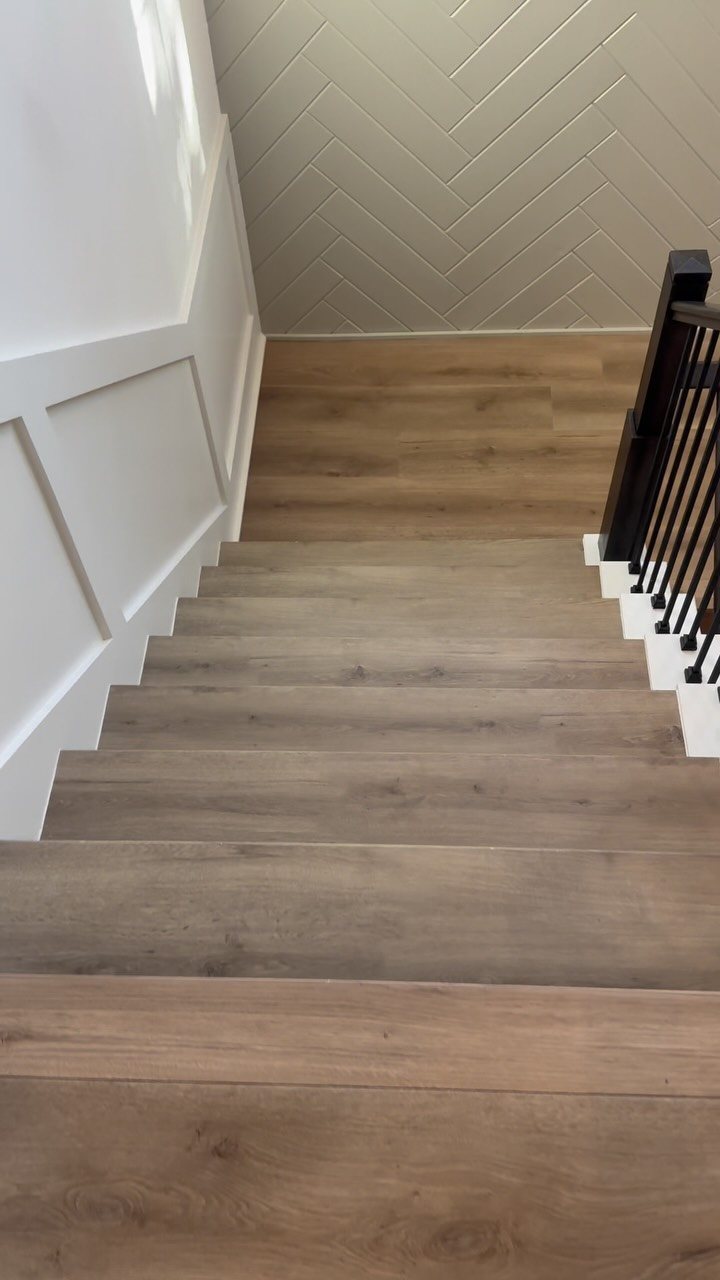 Wide plank light brown wood flooring installed on a staircase with white risers and black banister.