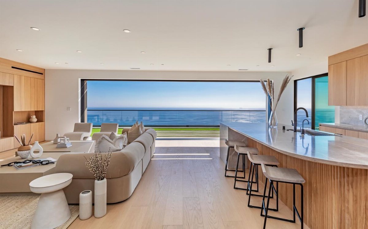 Modern living room with light wood flooring overlooking a vast blue ocean through large glass doors.