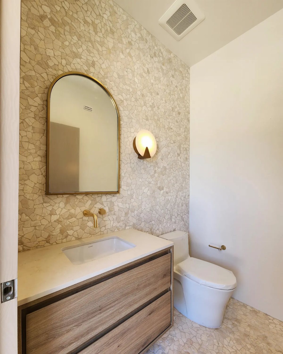 Small light-colored mosaic tiles covering a bathroom wall behind a wooden vanity and arched mirror.