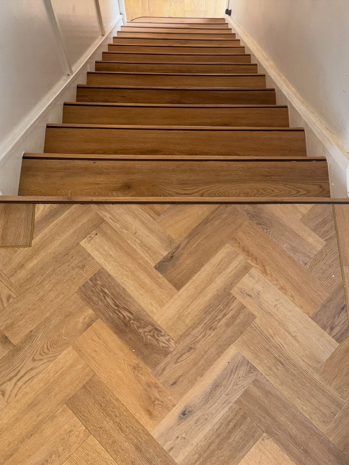 Wooden stairs leading upwards from a light oak herringbone pattern wood floor landing.