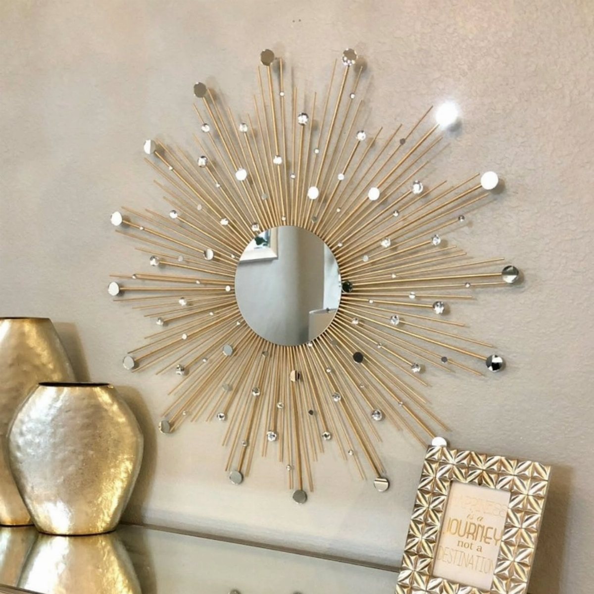 A gold sunburst mirror with crystal accents hangs on a beige wall above gold vases.
