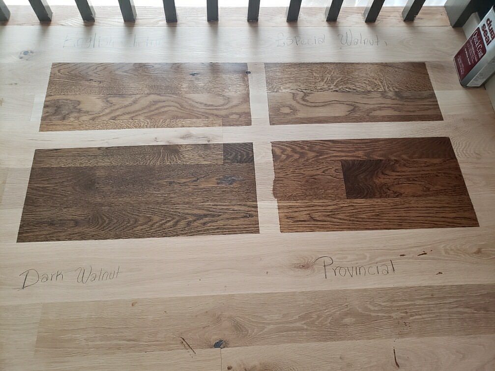 Four different wood stain samples applied to a light white oak floor with handwritten labels.