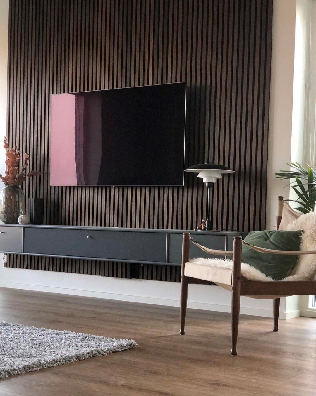 A television mounted on a dark smoked oak wood slat wall above a gray console.
